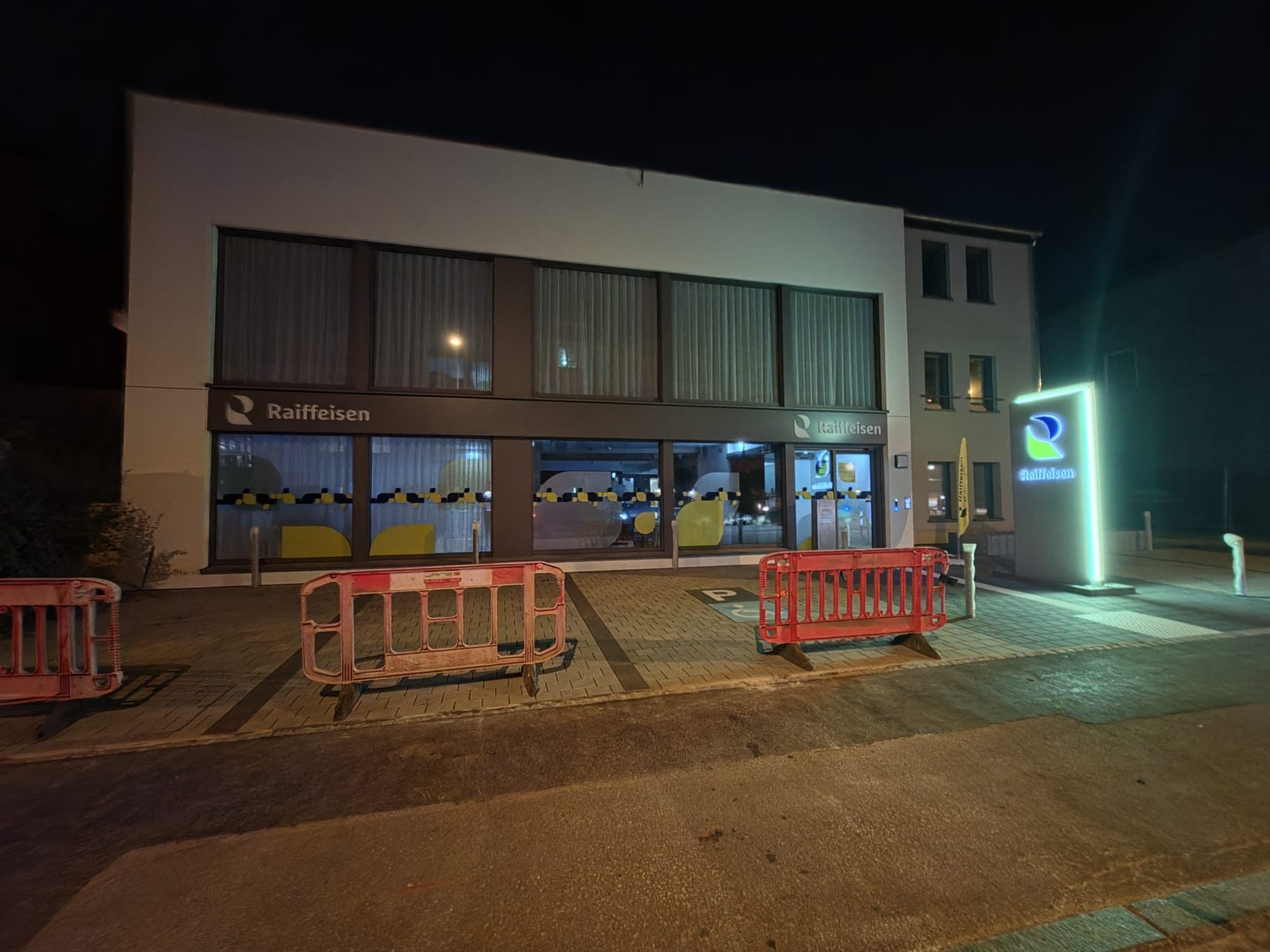 Raiffeisen-Filiale in Wiltz bei Nacht, beleuchtete Schaufenster und Totem